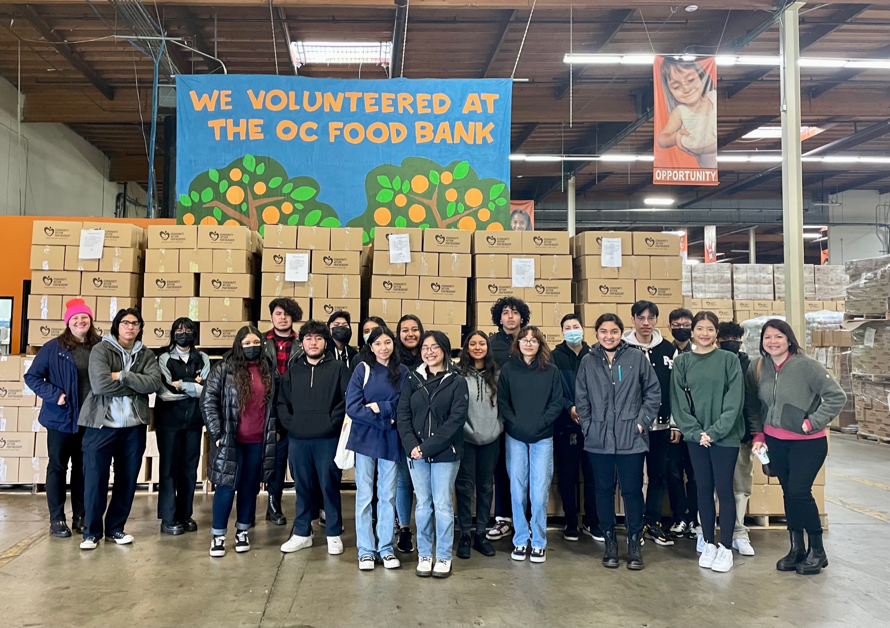 SAC ULink students and staff in a OC Food Bank warehouse posing in front of boxed food boxes and a sign that says "we volunteered at the OC Food Bank."